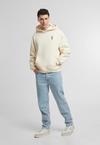 Kremfarget hettegenser med frontlomme og svart logo, parret med lyseblå jeans og hvite joggesko med beige detaljer.