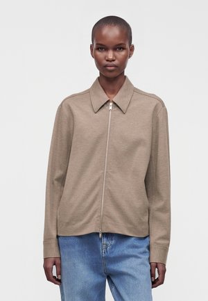 Persona con capelli corti che indossa una giacca beige con zip e colletto e jeans azzurri chiari, in piedi davanti a uno sfondo semplice.
