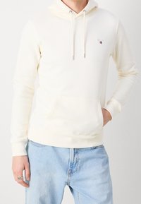 Sudadera de color crema claro hecha de una tela suave, cuenta con un bolsillo frontal, cordones ajustables y un pequeño logo en el pecho.