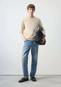 Joven hombre con suéter beige, vaqueros azules y zapatos negros sosteniendo una gran bolsa negra frente a un fondo claro y liso.