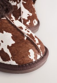 Brązowo-białe kapcie w cętki o miękkiej, futrzanej fakturze, z przeszywanym szwem i ciemnobrązową gumową podeszwą.