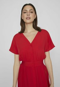 Czerwona sukienka z dekoltem w kształcie litery V, krótkimi rękawami i wcięciem w talii. Wykonana z lekkiego materiału, z guzikami umieszczonymi na przodzie.