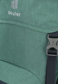 Zelený látkový batoh s čiernym nastaviteľným popruhom a šedým plastovým klipom, na ktorom je vyšité logo Deuter v bielej farbe.