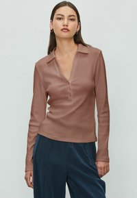 Una giovane donna con lunghi capelli scuri indossa una camicia colletto marrone con bottoni e pantaloni blu scuro, in piedi contro uno sfondo chiaro e semplice.