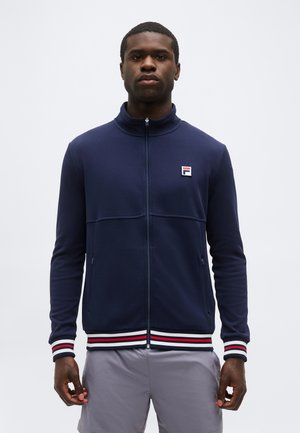 Mężczyzna w granatowej kurtce na zamek z logo Fila, z mankietami i dolnym ściągaczem w paski oraz w jasnoszarych szortach, stojący na tle jednolitej białej ściany.