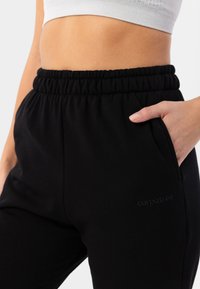 Melni sporta bikses ar elastīgu jostasvietu, sānu kabatām un diskretu zīmola logotipu, kas iepresēts uz kājas. Mīksta auduma tekstūra.