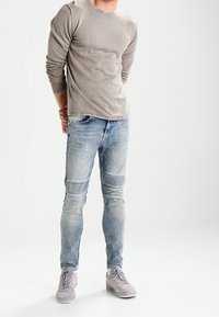 Homme portant une chemise à manches longues gris clair, un jean bleu décoloré avec des genoux en patch, et des baskets gris clair, se tenant les mains derrière le dos.