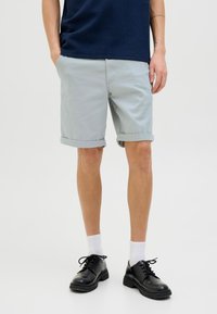 Helle graue Baumwollshorts mit umgeschlagenen Säumen, kombiniert mit schwarzen Schuhen und weißen Socken. Das Model trägt ein navyblaues Oberteil.