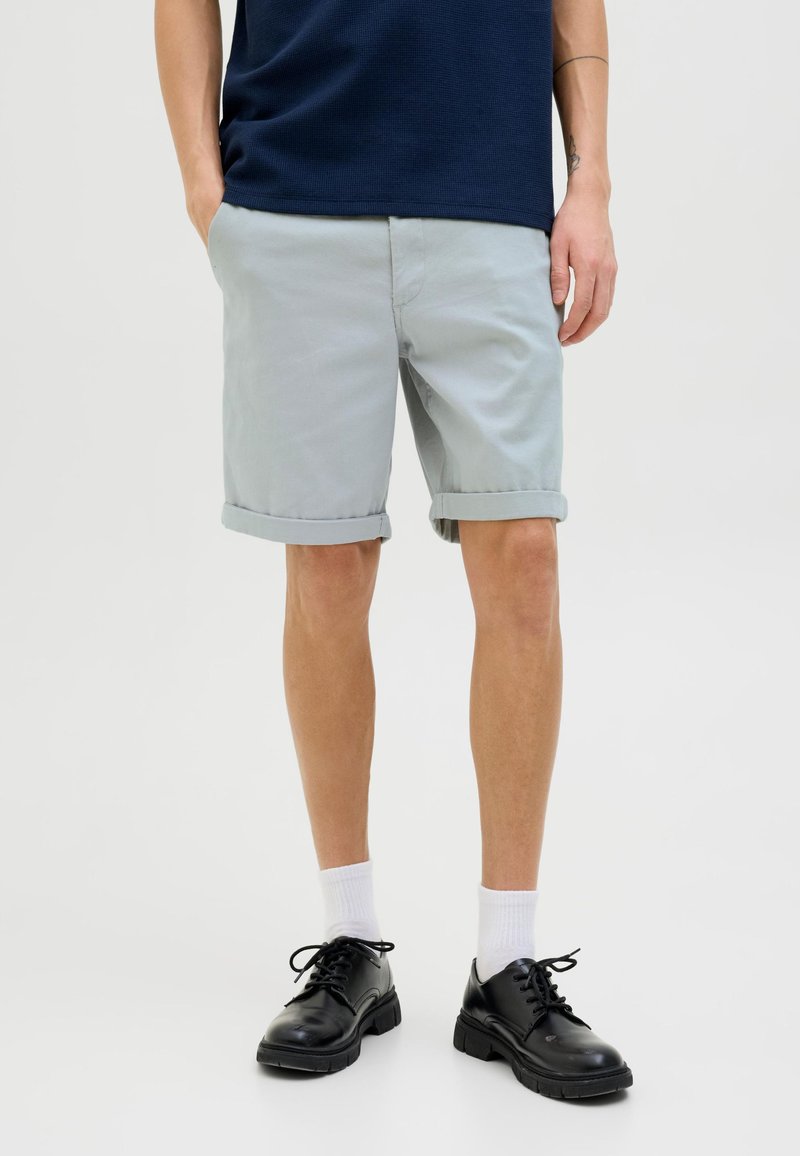 Helle graue Baumwollshorts mit umgeschlagenen Säumen, kombiniert mit schwarzen Schuhen und weißen Socken. Das Model trägt ein navyblaues Oberteil.