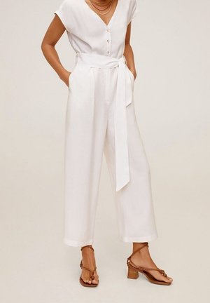 Frau trägt einen weißen ärmellosen Overall mit Taillengürtel und braunen Sandalen mit Absatz, steht mit den Händen in den Taschen.