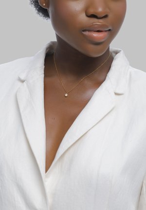 Frau trägt eine weiße Leinenbluse mit offenem Kragen, eine goldene Kette mit einem einzelnen kleinen Anhänger und einen kleinen Ohrstecker.