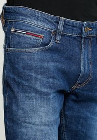 Jeans de mezclilla azul oscuro con corte clásico. Presenta un bolsillo frontal con un acento a rayas rojas y blancas, herrajes dorados y costuras visibles.