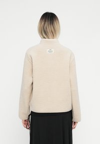  Άτομο με μαλλιά ξανθά μέχρι τον ώμο, ντυμένο με καφέ φλις μπουφάν και μακριά μαύρη φούστα, απεικονίζεται από πίσω σε λευκό φόντο.