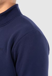 Maglione blu navy con colletto alto, realizzato in morbido cotone. Presenta una texture liscia e un design minimalista con branding discreto.