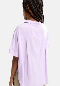 Jasnofioletowa koszula z krótkim rękawem w luźnym kroju, z kołnierzykiem i detalem w postaci zakładki na plecach. Gładki materiał o subtelnym połysku.