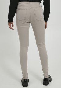 Vaqueros ajustados beige con una textura suave, diseño de cinco bolsillos y etiqueta de logo, combinados con calzado negro, vistos desde la parte de atrás.