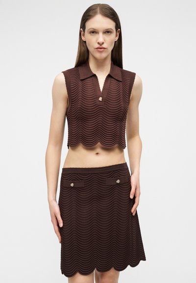 Femme portant un haut court marron texturé sans manches avec col, et une jupe assortie aux bords festonnés et aux rabats de poche boutonnés.