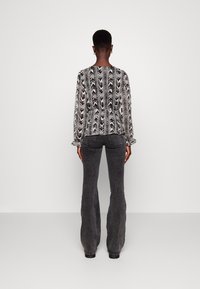 Persoon met kort haar die een zwart-wit gestreepte blouse met lange mouwen en donkergrijze flared jeans draagt, staand tegen een effen muur.