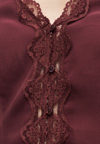 Ρουχισμός σε μπορντό με λουλουδάτη δαντέλα κατά μήκος της μπροστινής πλευράς με κουμπιά, εμφανίζοντας τρία κουμπιά και διαφανείς δαντελένιες λεπτομέρειες σε λείο ύφασμα.