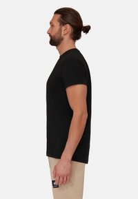 Schwarzes T-Shirt mit kurzen Ärmeln, normale Passform, Rundhalsausschnitt, aus Baumwollmaterial. Seitliche Ansicht, ergänzt durch beige Hosen mit einem Logopatch.