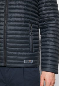 Vadderad mörkblå jacka med en zip-ficka, ribbad textur och en logotyp-plåster vid fållen. Strukturerad passform med kviltad design.