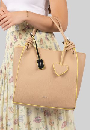 Woman holding beige leather handbag with yellow trim, knotted handles, black tag, and heart-shaped charm, wearing floral skirt and white top.