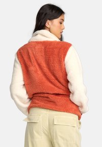 Fleece vest met een oranje rug en crèmekleurige mouwen. Beschikt over een zachte, gestructureerde stof, een kraag en een open ontwerp voor gemakkelijke layering.