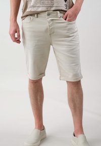 Shorts en denim beige avec ourlets roulés, cinq poches, délavage clair et fermeture à bouton. Portés avec des chaussures à enfiler grises. Surface texturée plate.