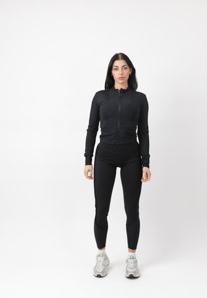 Donna in piedi che indossa una giacca nera con cerniera e leggings neri con sneakers bianche su sfondo bianco uniforme.