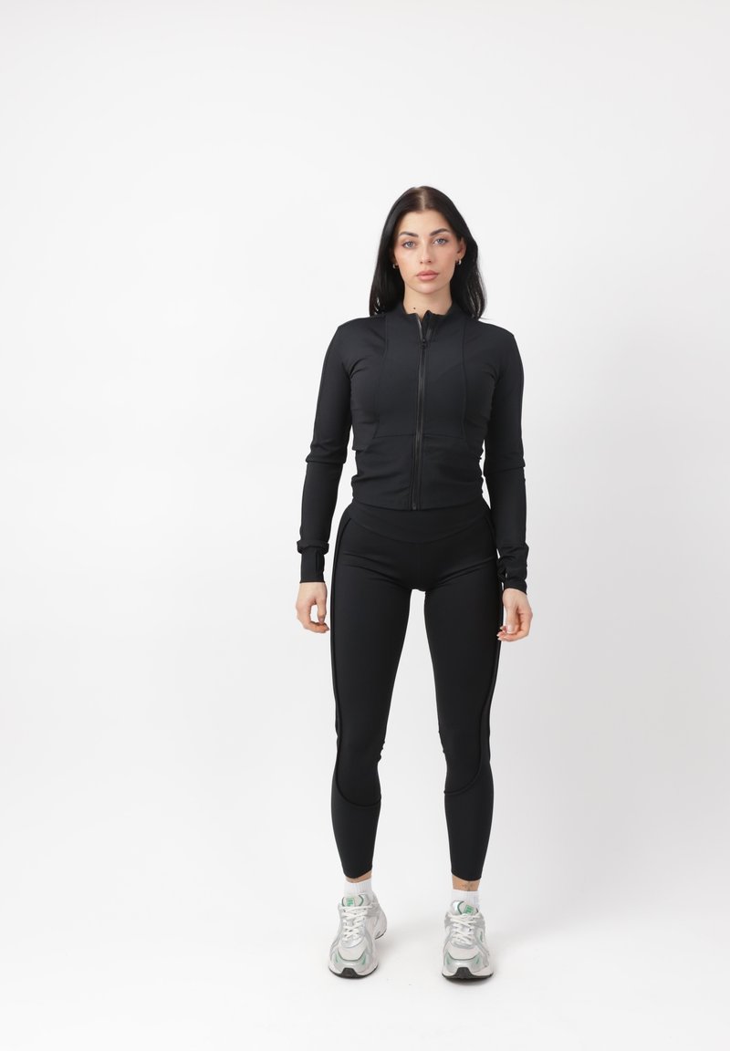 Donna in piedi che indossa una giacca nera con cerniera e leggings neri con sneakers bianche su sfondo bianco uniforme.