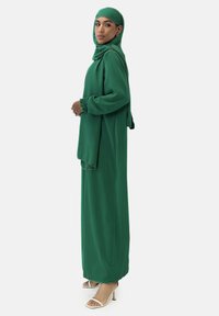 Grünes Kleid mit langen Ärmeln und passendem Hijab, mit fließendem Design und gerafften Bündchen. Getragen mit beigen Sandalen mit hohem Absatz.