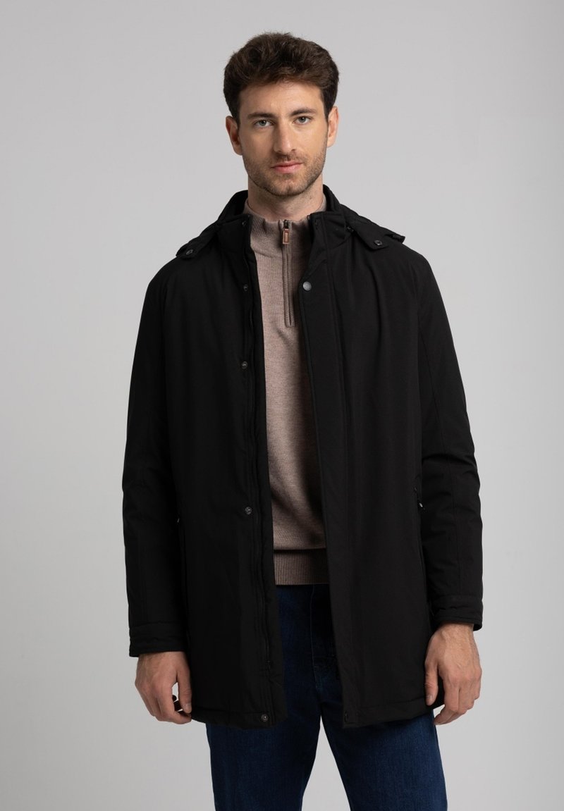 Hombre vestido con un abrigo negro con cremallera sobre un suéter beige de media cremallera y jeans azules, de pie frente a un fondo gris liso.