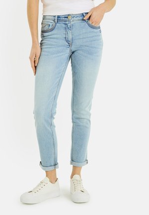 Femme portant un jean bleu clair retroussé, des baskets blanches et un haut blanc, debout avec une main dans la poche et une main détendue.