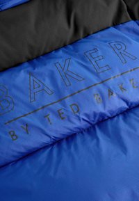Plava preklopna tkanina s sjajnom teksturom, s crnim panelom na vrhu i otisnutim natpisom "BAKER by TED BAKER" u crnoj boji.