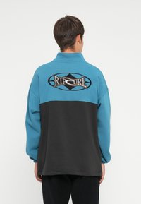 Blå og svart genser med høy krage, med et brodert "Rip Curl"-logo på ryggen og elastiske mansjetter.