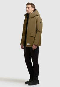 Khaki geïsoleerde jas met een hoge kraag en grote voorzak, gecombineerd met zwarte broek en laarzen, met een zwart logo-embleem.