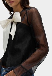 Blusa negra transparente con mangas largas y texturizadas, y un gran lazo blanco en el escote. Llevada sobre una camiseta negra, combinada con jeans azules.