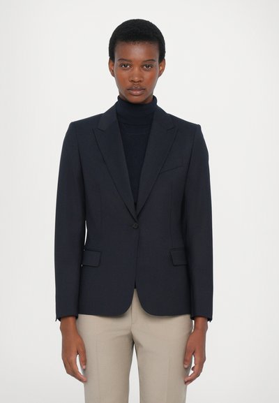 Blazer blu navy su misura con revers a punta, chiusura con un bottone e due tasche frontali, abbinato a un dolcevita nero. Tessuto con una texture liscia.