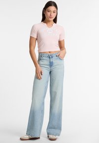 Heledaid roosa lühikeste varrukatega T-särk, millel on "Guess Jeans California" logo, koos laiemate siniste denimiste teksadega, millel on heledam pesu ja kõrge vöökoht.
