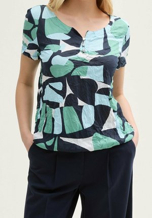 Femme portant un chemisier à manches courtes avec des formes abstraites bleues, vertes, noires et blanches, assorti à un pantalon foncé, mains dans les poches.