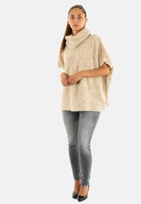 Beige gebreide poncho met een hoge kraag, zachte textuur, losse pasvorm en korte mouwen, gecombineerd met slim fit grijze jeans en zwarte hakken.