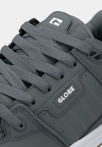 Grijze sneaker met synthetisch bovenwerk, brede platte veters en een gestructureerde afwerking. Bevat een wit logo en subtiele stikselaccenten aan de zijkant.