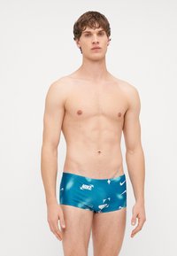 Calções de banho turquesa com um acabamento brilhante, apresentando uma mistura de logótipos brancos da Nike e padrões abstratos. Corte padrão com uma cinta elástica.