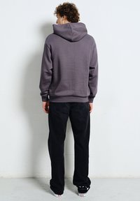 Osoba o kręconych włosach ubrana w szarą bluzę z kapturem, czarne dżinsy i czarne trampki stoi twarzą do gładkiej białej ściany.