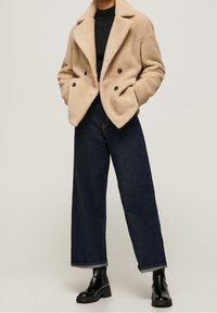 Osoba w beżowym płaszczu teddy, czarnym swetrze ze splotem, ciemnych szerokich dżinsach i czarnych błyszczących botkach za kostkę na jednolitym tle.