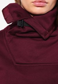 Sudadera burdeos con cuello alto, que presenta un diseño de solapa y un detalle de hebilla negra en el cuello. Textura suave y estilo minimalista.