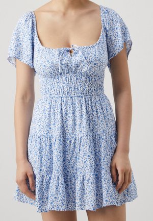 Frau trägt ein kurzes blau-weißes Blumenmusterkleid mit flatternden Ärmeln, einem quadratischen Ausschnitt und gerafften Taillendetails.