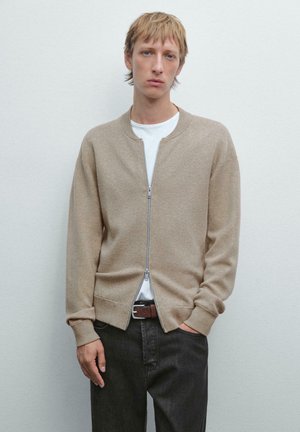 Beige gestrickte Bomberjacke mit einem durchgehenden Reißverschluss, gerippten Bündchen und zwei Seitentaschen, kombiniert mit einem weißen T-Shirt und dunklen Jeans.