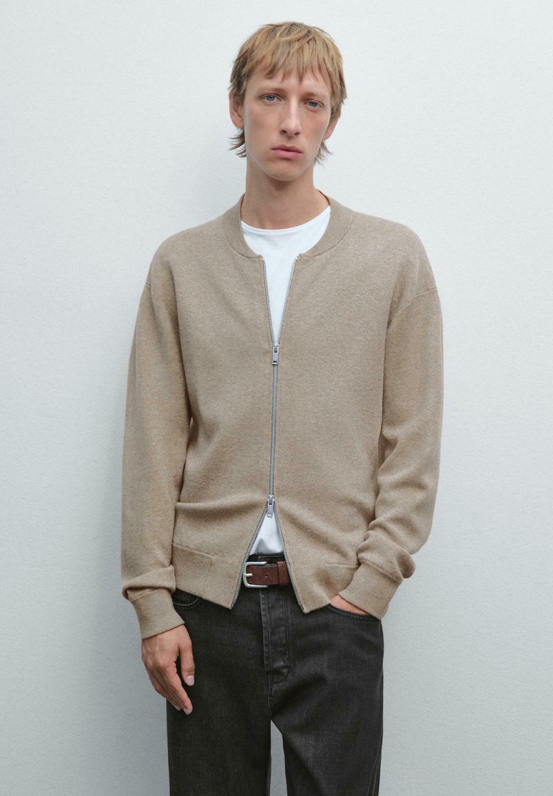 Giubbotto bomber beige lavorato a maglia con zip completa, polsini a coste e due tasche laterali, abbinato a una maglietta bianca e jeans scuri.