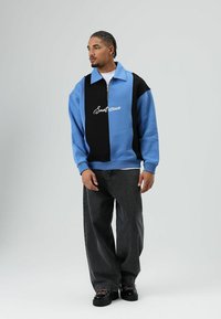 Sweat-shirt zippé bleu clair et noir avec grand col et logo brodé ; associé à un jean large gris foncé et des chaussures noires.